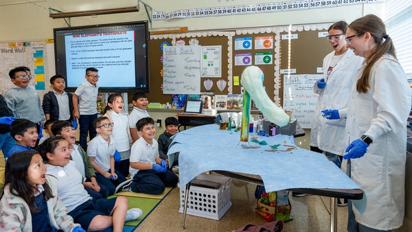 Children sitting on the floor of a classroom watch a science demo overseen by two lab coat wearing women.
