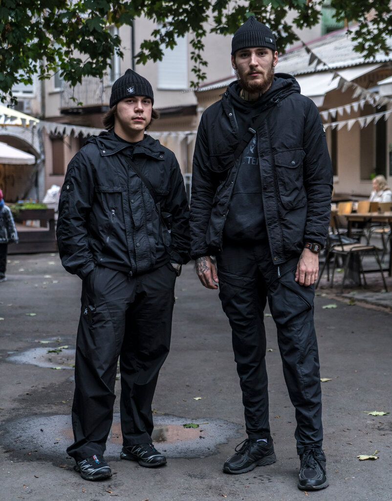 Two people wearing black outfits and caps stand on a street.