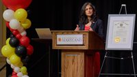 woman speaks at podium next to red, white, black and gold balloons