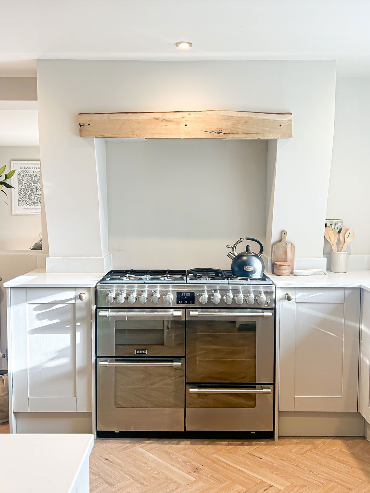 Crisp white walls, counters and cupboards bounce the sunlight around this beautiful kitchen. The gleaming stainless steel range awaits the next culinary creation, but for the blue kettle is ready to boil up water for a coffee. The wooden utensils tie in with the original pale wooden beam over the hob, cleverly hiding the extractor from view. Beyond the kitchen area, a beautiful arrangement of white flower and foliage gives us just a glimpse of the room beyond with its monochrome framed print on the wall.