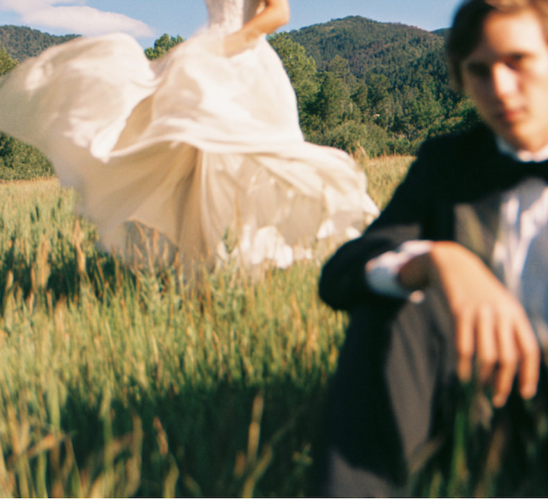 A bride in a flowing white dress twirls in a sunlit meadow, her motion creating a soft blur, while a man in a tuxedo sits in the foreground, slightly out of focus. Lush green grass and forested mountains fill the background under a blue sky with scattered clouds.