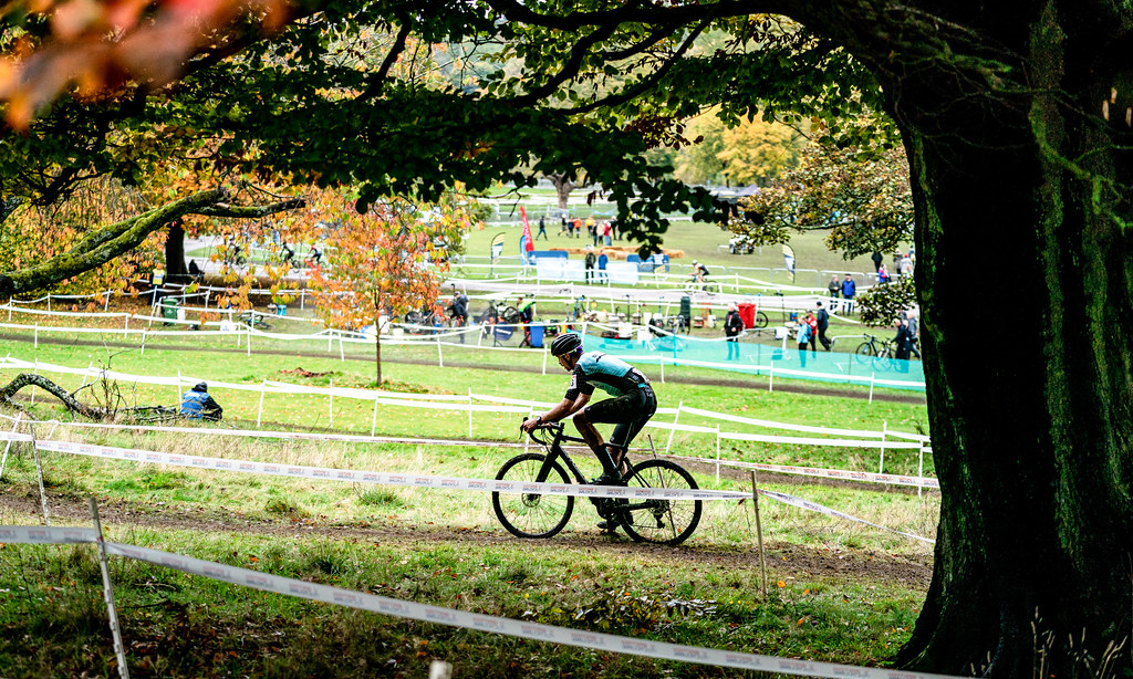 A cyclo-cross rider mid-shot during an event, with spectators and event infrastructure in the background.