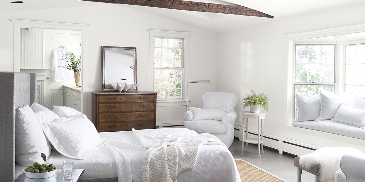 A cozy, white-painted bedroom with white bedding and chairs, a bay window with white cushions, an antique dresser, and a vaulted white ceiling with a dark brown wooden beam, looking into a white bathroom.