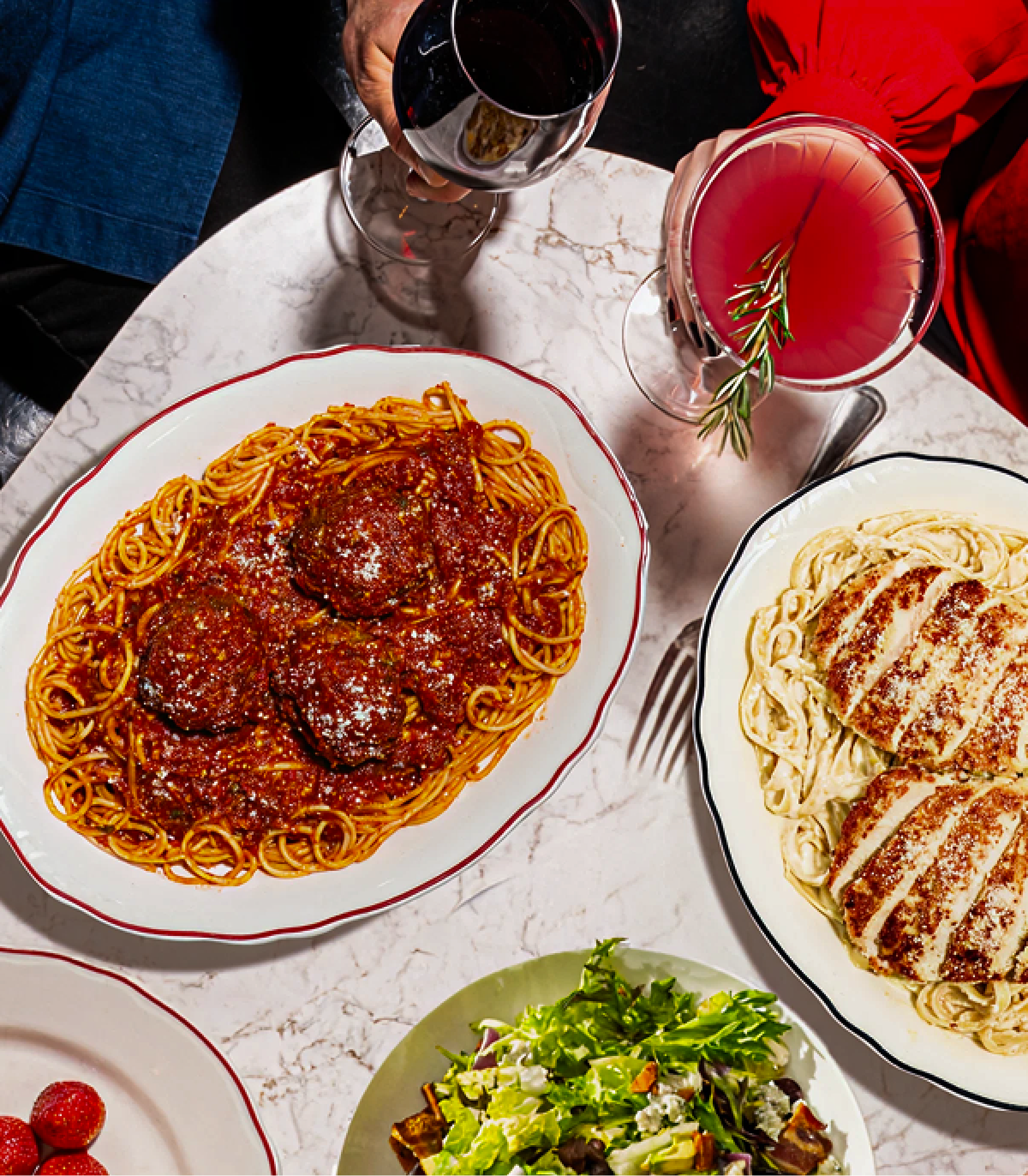 Overhead shot of a couple with drinks and a few Maggiano’s entrées, including Spaghetti & Meatballs, Chicken Fettuccine Alfredo, and Maggiano’s salad.