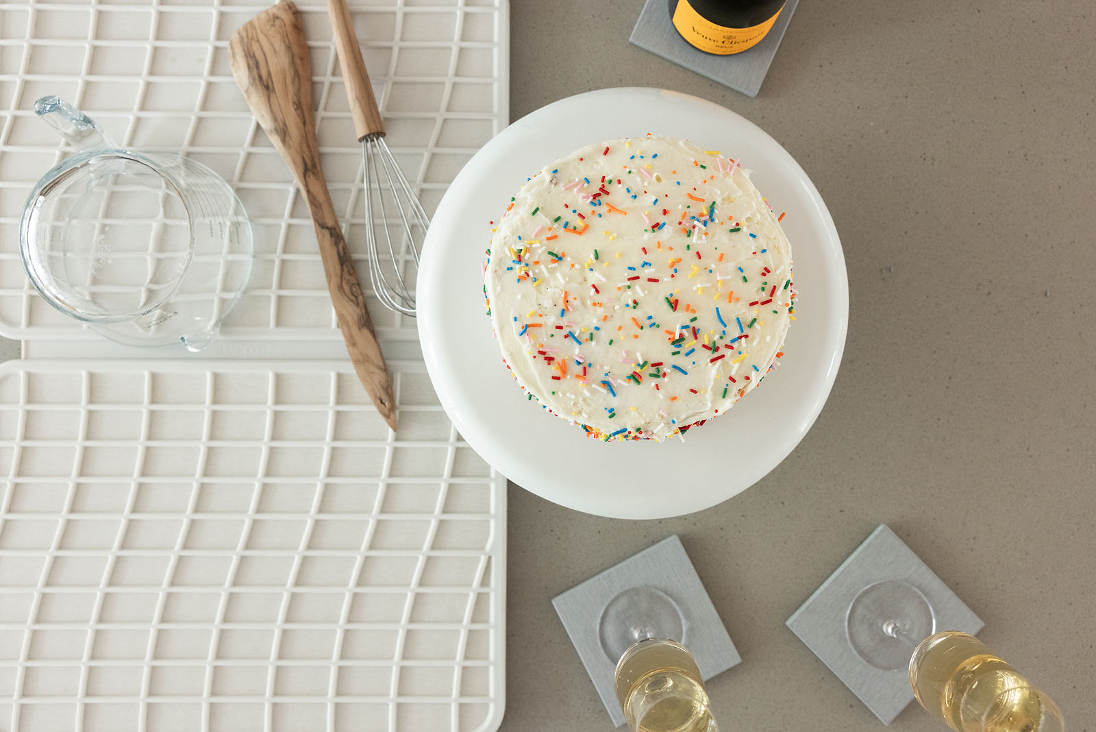 Overhead view of a frosted white cake with rainbow sprinkles on a white cake stand, styled on a kitchen countertop beside a white drying mat holding a glass measuring cup, wooden spatula, and whisk. Two wine glasses rest on light gray stone coasters nearby.