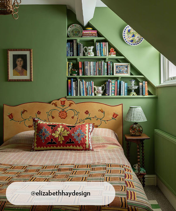 A characterful soft green bedroom featuring a hand-painted floral headboard, patterned textiles, and a colourful mix of books and ceramics on built-in shelves. The eclectic layers and warm detailing give the room a cosy, whimsical feel.