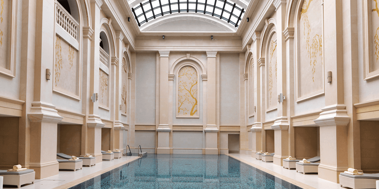A rectangular pool in a Grecian-style room made of cream-colored stone, featuring a high ceiling.