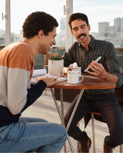 Two men sit at a rooftop patio table looking at real estate on the Zillow app.