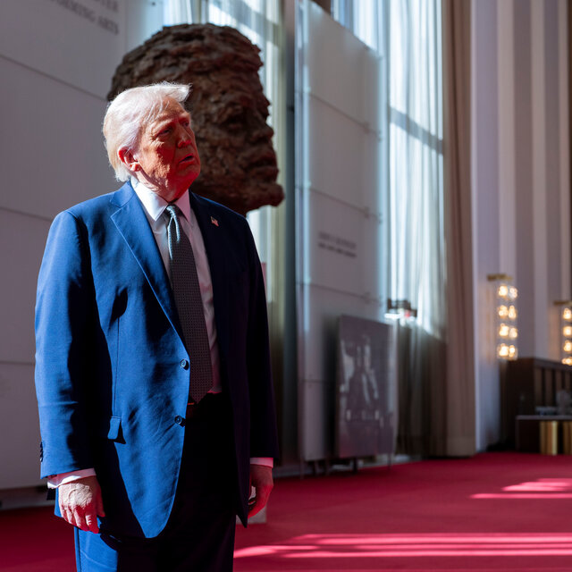 President Trump, in a blue suit, looks upward while standing in front of a large bust of President Kennedy’s head.